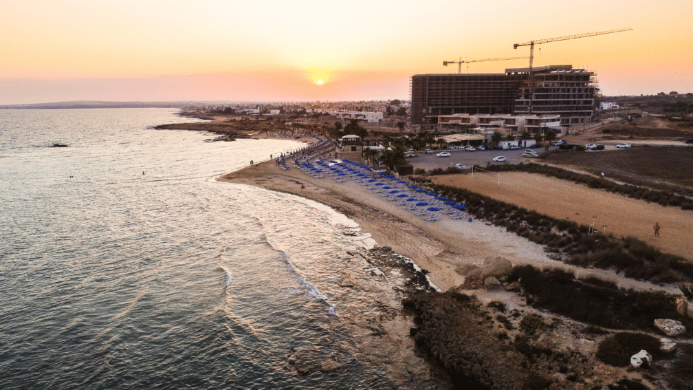 A landscape photo of the sea, surrounded by a beach. Buildings are in the background and the sunset.